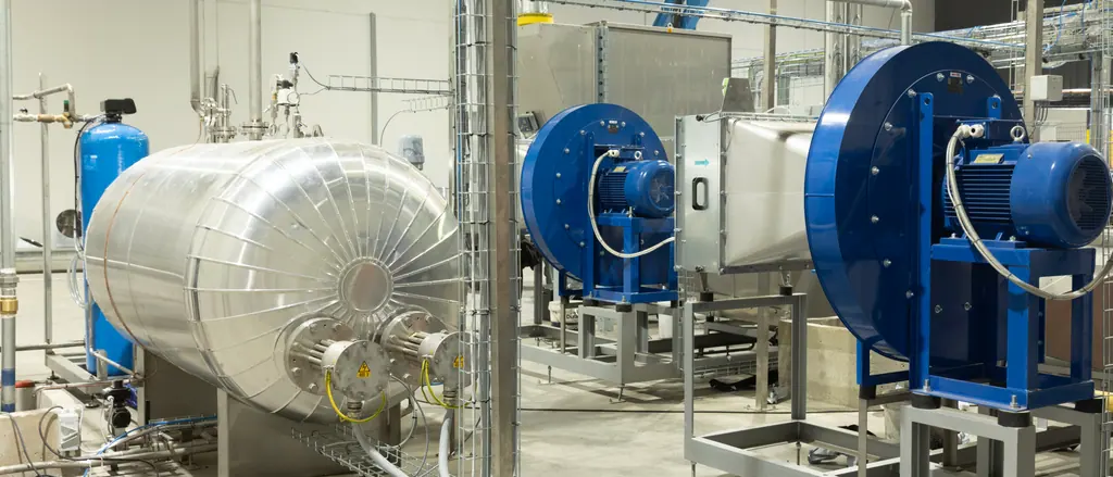 The Temong plant floor: an insulated steam vessel on the left, two blue industrial blowers on steel frames to the right, overhead pipework running the length of the hall.