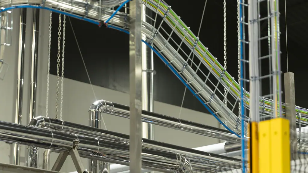 Looking up at the plant services: galvanised cable trays carrying colour-coded conductors, polished steam lines and suspension chains against a dark roof.
